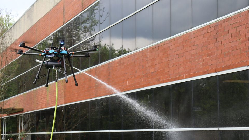 A tethered cleaning drone washes the glass and brick of an office building.